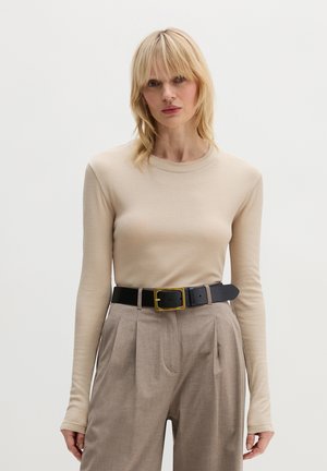 Blonde Frau trägt ein beiges, langärmliges, eng anliegendes Oberteil, das in eine hoch taillierte, hellbraune, plissierte Hose mit schwarzem Gürtel gesteckt ist.
