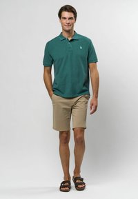 Tyrkysové polo tričko s krátkymi rukávmi, svetlohnedé šortky a hnedé sandále. Polo tričko má malé logo na hrudi.