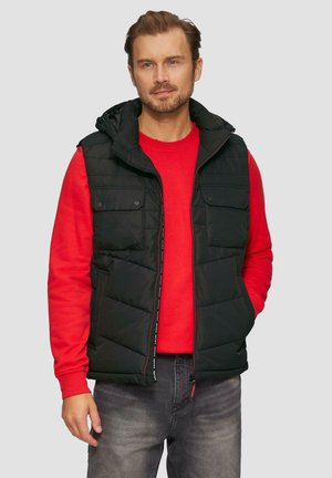 Zwarte gewatteerde vest met een capuchon, voorzien van een ritssluiting aan de voorkant, twee borstzakken en een quilted ontwerp, gedragen over een rode sweatshirt.