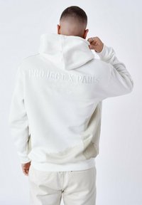 Sudadera blanca con un acabado texturizado, presentada con un logotipo en relieve en la parte trasera y un diseño salpicado de pintura en beige claro.