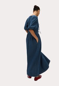 Denim maxi jurk in donkerblauw met pofmouwen, een cinched taille, zijzakken en een vloeiende rok. Heeft een striksluiting aan de achterkant.