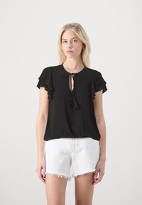 Svart blouson-topp med korte volang-ermer, rund hals og knytedetalj foran, paret med hvite shorts med frynsete kant.