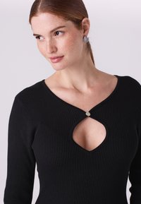 Čierny rebrovaný top s výrezom v tvare kľúčovej dierky na výstrihu, zdobený okrúhlym kovovým gombíkom a dlhými rukávmi.