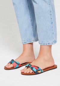 Sandali multicolore con parte superiore in tessuto floreale, caratterizzati da un design annodato e suola in gomma marrone. Indossati con jeans azzurri cropped.