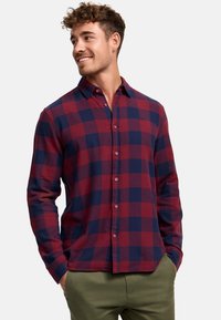 Camisa de cuadros roja y azul marino con botones, hecha de una tela de textura suave; cuenta con un collar y mangas largas, combinada con pantalones verde oliva.