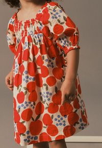 Vestido con un estampado de manzanas rojas y arándanos sobre un fondo beige. Las mangas cortas abullonadas y el escote fruncido añaden textura y detalle.