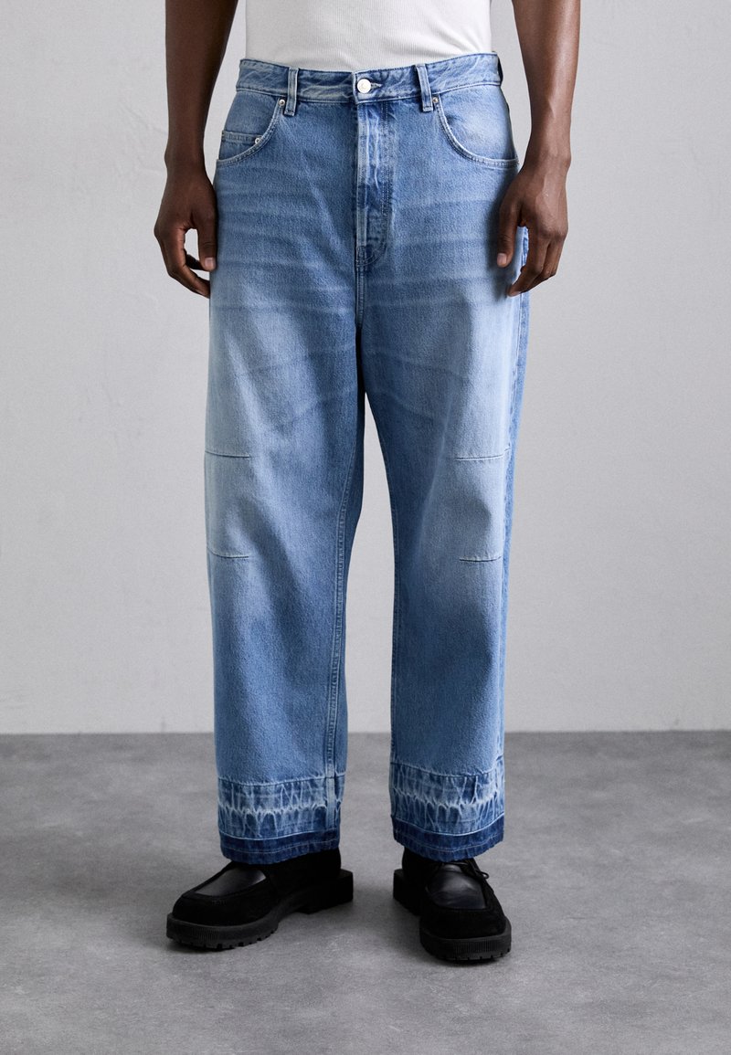 Jeans large en denim bleu clair avec une finition délavée, featuring des coutures à double surpiqûre et un motif tie-dye unique au niveau des ourlets.