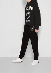 Schwarzer, kürzer geschnittener Hoodie mit weißem Logo, kombiniert mit schwarzen Sweatpants mit einem Bärenmotiv aus Strasssteinen. Weiße Sneakers runden das Outfit ab.