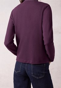 Haut à manches longues violet foncé avec une finition texturée, présentant un col rond et un ourlet droit, associé à un jean en denim bleu foncé.