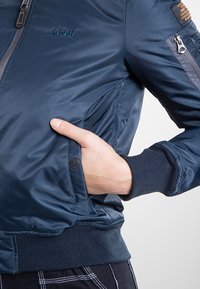 Granatowa kurtka bomberka z gładkiego nylonu z przednim zamkiem błyskawicznym, ściągaczami na mankietach i boczną kieszenią. Posiada logo i dodatkową kieszeń na zamek na rękawie.