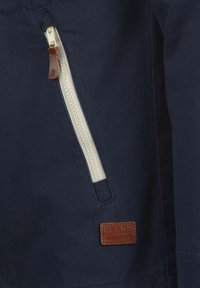Section de veste marine avec une fermeture éclair blanche contrastante munie d'une tirette en cuir, et une étiquette de marque en cuir marron avec texte embossé.