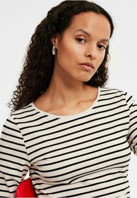 Gestreepte lange mouw top in crème en zwart, met een ronde halslijn en zachte stof met een gladde textuur. Model draagt zilveren hoepel oorbellen.