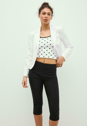 Jonge vrouw met krullend haar die een witte blazer, een witte cropped top met zwarte stippen en zwarte cropped leggings draagt, staand tegen een lichte achtergrond.