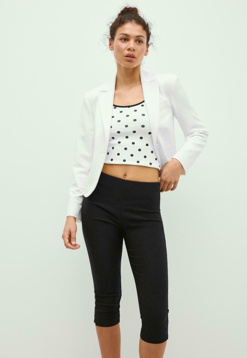 Jeune femme aux cheveux bouclés portant un blazer blanc, un crop top blanc à pois noirs et des leggings courts noirs, debout devant un fond clair.