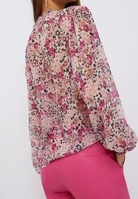 Blusa floral de tejido ligero y transparente con patrones en rosa y marrón, mangas abullonadas y escote fruncido. Combinada con pantalones rosa.