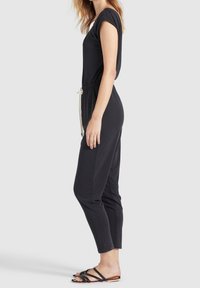 Zwarte jumpsuit met korte mouwen, getailleerde taille en trekkoorddetails. Glad, rekbaar materiaal; taps toelopende pijpen. Draag met platte zwarte sandalen.