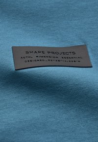 Tela azul con una textura suave presenta una etiqueta rectangular negra. La etiqueta tiene texto en relieve: "SHAPE PROJECTS," "AUTH. DIMENSION ESSENTIAL," y coordenadas.
