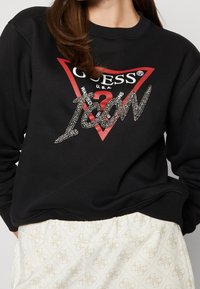 Sudadera negra con logo de triángulo rojo, texto "GUESS" y adornos plateados. Puños y dobladillo acanalados, combinada con parte inferior ligera y estampada.