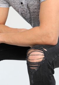 Jeans negros desgastados con roturas en las rodillas, con bordes deshilachados y un ajuste slim. La persona lleva una camisa polo de textura gris.