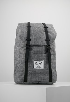 Sac à dos en tissu gris avec des sangles à boucle noire et une poche avant présentant une étiquette rectangulaire Herschel Supply Co., affiché sur une surface blanche.