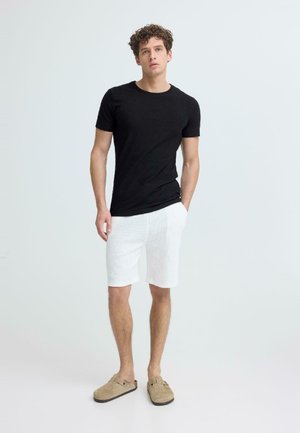 Mladi moški stoji obrnjeno naprej, oblečen v črno majico s kratkimi rokavi, bele kratke hlače in bež čevlje brez zavezovanja, na svetlem ozadju.