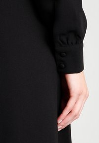 Černé šaty s dlouhým rukávem, které mají knoflíkové manžety, hladkou texturu a na zápěstí nařasenou látku, zdůrazňující minimalistický design.