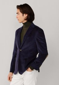 Blazer de pana en color marino con cierre de botón único, solapas de muesca y textura sutil, combinado con un jersey de cuello alto verde oscuro y pantalones blancos.