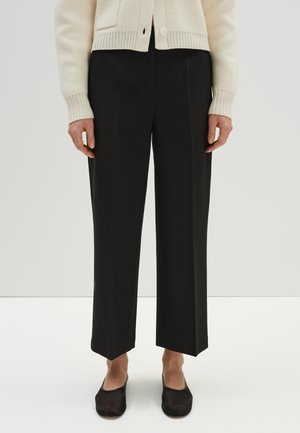 Schwarze weit geschnittene Hose mit taillierter Passform, geradeem Saum und vorderer Bügelfalte, kombiniert mit schwarzen Slippers.