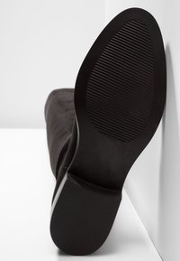 Semelle de bottine noire avec dessin de semelle crantée et petit talon bloc reposant contre une surface blanche.