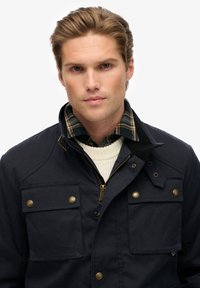 Marineblauwe jas van duurzaam materiaal, met twee grote voorzakken met gouden hardware, een ritssluiting en een zwarte kraag over een crèmekleurige trui.