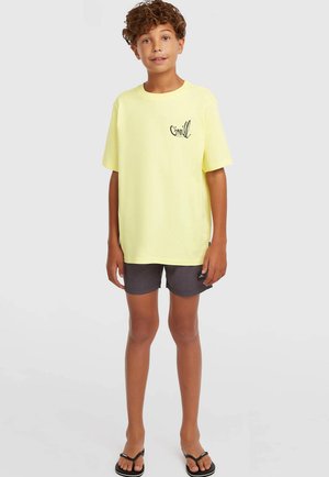 Żółta bawełniana koszulka z okrągłym dekoltem i czarnym logo na piersi, zestawiona z ciemnoszarymi szortami i czarnymi klapkami.