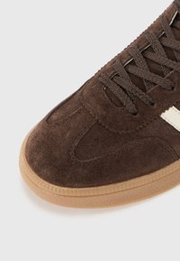 Sneaker in suede marrone con lacci scuri, suola in gomma e dettagli bianchi. Presenta una punta arrotondata e una parte superiore testurizzata per presa e comfort.