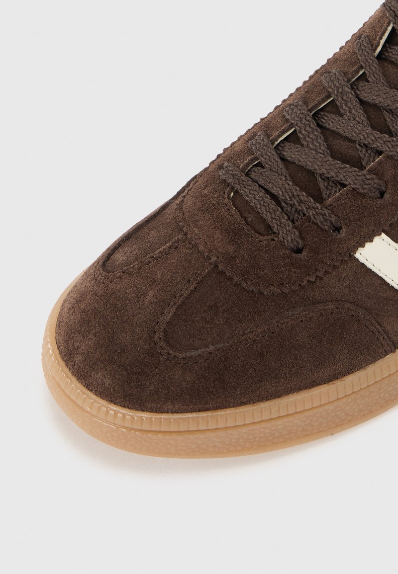 Braune Wildleder-Sneaker mit dunklen Schnürsenkeln, Gummisohle und weißen Akzenten. Mit abgerundetem Zehenbereich und texturiertem Obermaterial für Grip und Komfort.