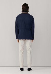 Chaqueta impermeable azul marino con cuello abotonado y detalle de costura diagonal, combinada con pantalones de color claro y zapatos oscuros, mostrada desde atrás.