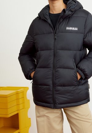 Person wearing a black puffer jacket with a hood and zipped pockets, standing with hands in pockets against a plain background.