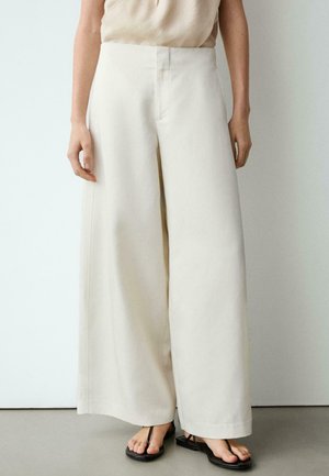 Frau trägt hoch taillierte, weit geschnittene, off-white Hose kombiniert mit beigem Oberteil und schwarzen Zehensandalen und steht vor einer schlichten Wand.