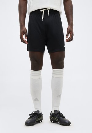 Männlicher Athlet, der schwarze Shorts mit Kordelzug, weiße knielange Socken und schwarze Stollenschuhe mit gelben Akzenten trägt, steht vor einem schlichten Hintergrund.