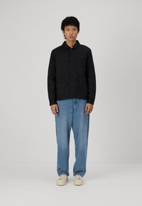 Svart knappete jakke med krage, frontlommer og avslappet passform, paret med lyseblå jeans og hvite joggesko.
