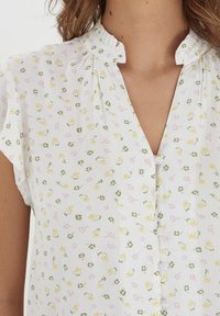 Weiße Bluse mit einem dezenten strukturierten Muster und gelben sowie pinken Blumenmotiven. Sie hat kurze Ärmel und einen V-Ausschnitt mit Knöpfen.