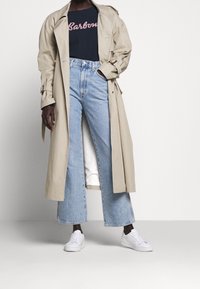 Beige trenchcoat over een marineblauwe t-shirt met roze letters, gecombineerd met hooggetailleerde lichtblauwe wijde jeans en witte sneakers.