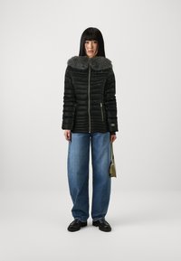 Svart pufferjakke med grå pelskrage, glidelås, quiltet design, sammen med avslappet passform blå jeans og kraftige svarte sko.