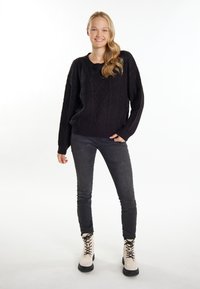 Schwarzer Strickpullover mit strukturierten Rautenmuster, kombiniert mit dunklen Jeans und cremefarbenen Schnürstiefeln. Weißer Hintergrund.