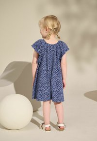 Robe bleu à motifs avec des manches flottantes, coupe décontractée, longueur genoux. Sandales blanches avec bride à la cheville. Arrière-plan neutre et sculpture ronde.