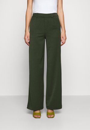 Femme portant un pantalon taille haute à jambes larges vert foncé, associé à des talons verts ouverts et un top blanc sans manches, debout devant un fond uni.