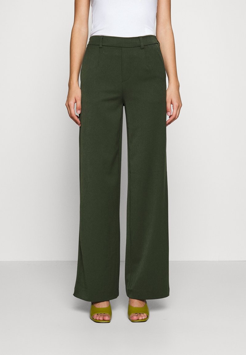 Femme portant un pantalon taille haute à jambes larges vert foncé, associé à des talons verts ouverts et un top blanc sans manches, debout devant un fond uni.