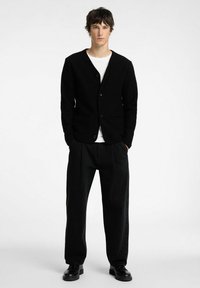 Mustra tekstuuriga must kardigan, millel on V-kaelus, nööpide sulgemine ja kaks esist kotti, koos vabalt langevate mustade pükstega.
