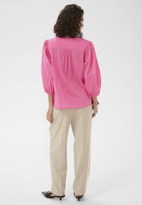 Blouse rose avec manches bouffantes, détail plissé au dos et encolure arrondie, associée à un pantalon droit couleur crème et des talons noirs.