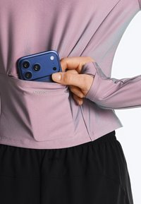 Ruka vkládající modrý chytrý telefon do malé zadní kapsy růžově fialového dlouhého topu s průstřely na palce a černých kalhot.