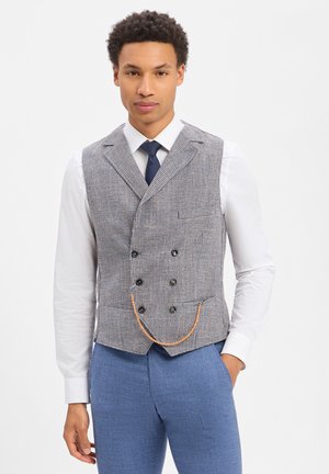 Mann trägt graue, doppelreihige Weste mit Kette, weißes Hemd, marineblauen Krawatte und blaue Hose, steht mit einer Hand in der Tasche.