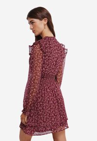 Robe floquée bordeaux avec des manches longues transparentes, des détails de manches volantes et une taille smockée. Elle présente une jupe à plusieurs couches et de petits motifs floraux.